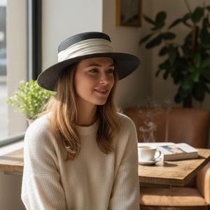 May include: A black and white fedora-style hat with a cream-colored band. The hat is worn by a person wearing a cream-colored sweater. The background includes a wooden table, a coffee cup, and a plant.