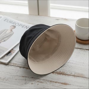 May include: A black bucket hat with a beige interior. The hat is made of a soft material and is designed to be reversible. The hat is on a light-coloured wooden surface.