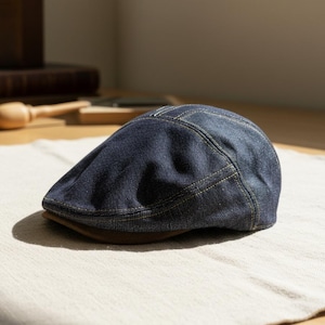 May include: A dark blue denim flat cap with a brown brim. The cap has yellow stitching details and is resting on a light-colored fabric. The hat is a classic style.