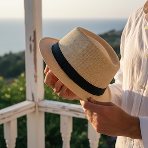 May include: A beige fedora hat with a black band. The hat is held by a person. The hat has a structured crown and a slightly curved brim. The background shows a coastal view with a white railing.