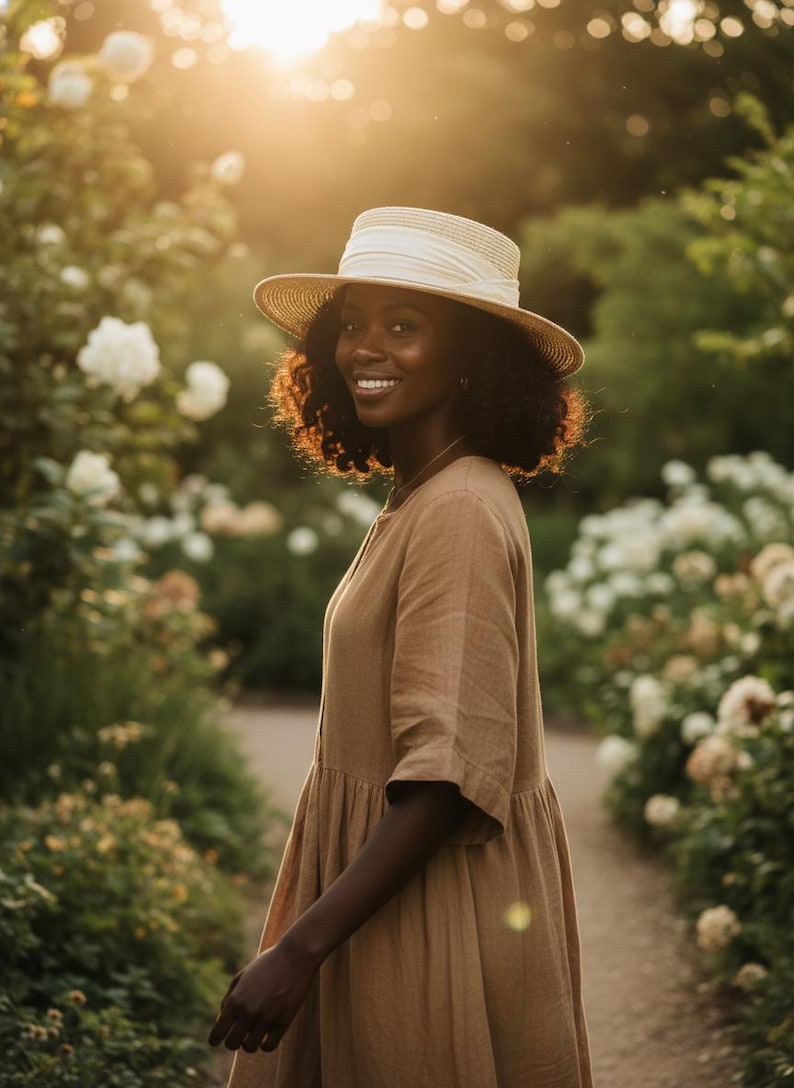 May include: A woman wearing a tan dress and a straw hat with a white band smiles in a garden. The dress has short sleeves and a flowing skirt. The hat has a wide brim. The background features white flowers and greenery.