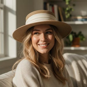 May include: A straw hat with a wide brim and a cream-colored band. The hat is tan and cream-colored, and is worn by a person with blonde hair. The person is wearing a cream-colored sweater.