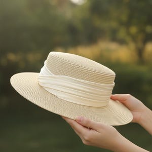 Könnte beinhalten: Ein beigefarbener Strohhut mit breiter Krempe und zylindrischer Krone. Der Hut hat ein cremefarbenes Band um die Krone gewickelt. Der Hut wird vor einem verschwommenen grünen Hintergrund gehalten.