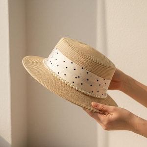 May include: A beige straw hat with a flat top and brim. The hat features a white band with black polka dots and a row of faux pearls. The hat is held by a person's hands.