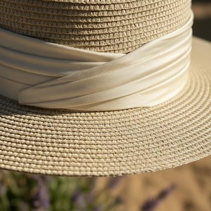 May include: A close-up of a straw hat with a wide brim and a cream-colored satin band. The hat is made of woven straw and has a classic, elegant design. The band is wrapped around the crown of the hat.