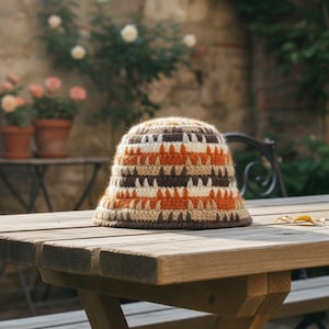 May include: A crocheted bucket hat in brown, orange, and cream tones. The hat is placed on a wooden table outside. The design features horizontal stripes. The hat is a stylish accessory.