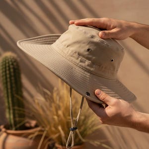Puede incluir: Sombrero para el sol de color beige con ala ancha y correa para la barbilla. El sombrero tiene un diseño estructurado con una superficie texturizada y ojales de ventilación. Lo sostienen dos manos, mostrando su forma y características. Accesorio práctico.