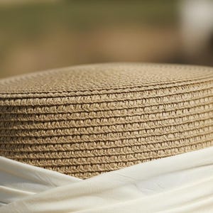 May include: Close-up of a woven straw hat with a cream-colored fabric band. The hat has a round crown and a wide brim, perfect for sun protection. The natural beige color of the straw contrasts with the white fabric.