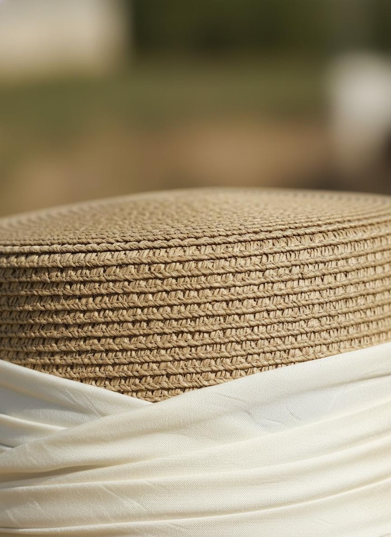 May include: Close-up of a woven straw hat with a cream-colored fabric band. The hat has a round crown and a wide brim, perfect for sun protection. The natural beige color of the straw contrasts with the white fabric.