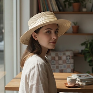 May include: A beige straw hat with a cream-colored band. The hat has a wide brim and a rounded crown. The hat is worn by a person in a light-colored blouse. The background includes a wooden table, books, and a coffee cup.