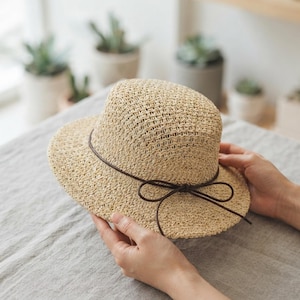 May include: A light beige straw hat with a rounded crown and a medium-width brim. The hat features a thin, dark brown cord tied into a simple bow around the base of the crown, providing a subtle contrast to the woven texture of the straw material.