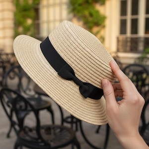 May include: A beige straw hat with a black ribbon and bow. The hat has a wide brim and a structured crown. The black ribbon is wrapped around the base of the crown and tied into a bow on the side.