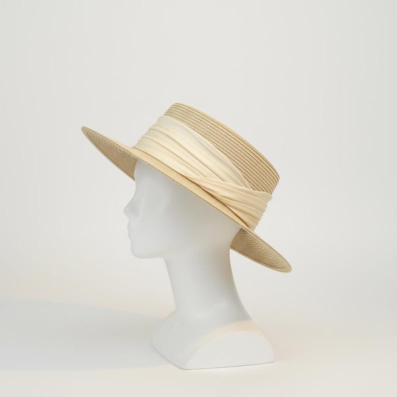 May include: A straw hat with a wide brim and a cream-colored band. The hat is displayed on a white mannequin head. The hat's brim measures approximately 3 inches.