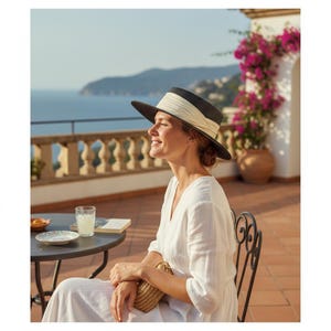 May include: A woman wearing a black and white sun hat with a cream-colored band, sitting outdoors. She is wearing a white dress and holding a woven clutch. A table with a drink and a book is in front of her.