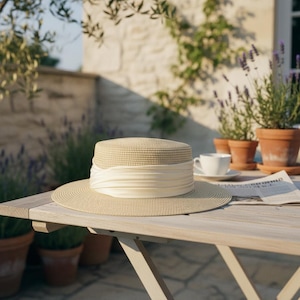 May include: A beige straw hat with a wide brim and a cream-colored ribbon band. The hat sits on a wooden table, with a teacup and saucer, and potted plants in the background. The hat is a classic accessory for sun protection.