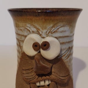 Könnte beinhalten: Eine handgefertigte Keramiktasse mit einem geformten Gesichtsdesign. Die Tasse hat ein braunes und beiges Farbschema mit großen weißen Augen und einem detaillierten braunen Gesicht. Ein einzigartiges Keramikstück.