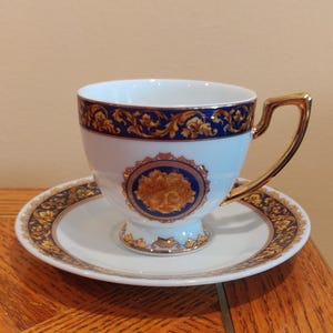 May include: A white teacup and saucer set with gold accents and a blue border. The teacup has a gold handle and a decorative emblem on the front. The saucer and cup feature a gold and blue ornate design.