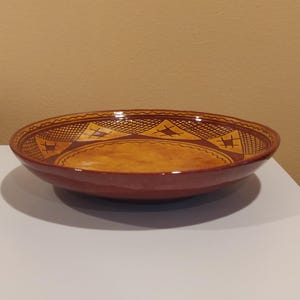 May include: A round, ceramic bowl with a wide rim and a shallow center. The bowl is primarily reddish-brown with a yellow-orange interior. The rim features a repeating geometric pattern in a darker brown color.