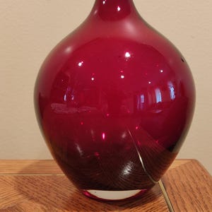 Peut inclure: Un vase en verre rouge foncé avec un corps arrondi et un col étroit. Le vase a une finition brillante et repose sur une surface en bois. Le vase est un objet décoratif.