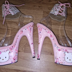 May include: A pair of pink platform high heels featuring a Hello Kitty design. The heels have clear straps, rhinestone detailing, and decorative embellishments. The platform base displays a Hello Kitty face with a pink bow.