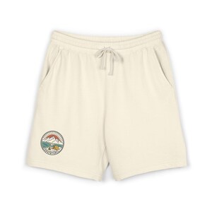 May include: Off-white sweat shorts featuring an elastic waistband with a drawstring. A circular graphic on the lower left leg depicts a mountain landscape, a tent, and a campfire with the word ADVENTURE printed in black along the bottom edge.