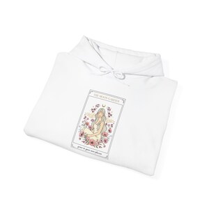 May include: White hoodie featuring a tarot card design. The card depicts a woman with long hair, surrounded by flowers, with the text "THE MOON GARDEN" and "grow in your own phases". The design is in soft pastel colors.