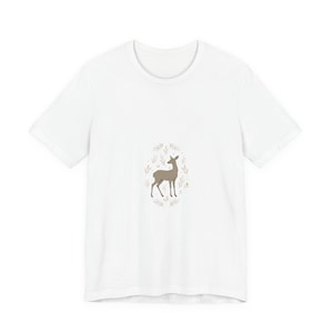 May include: White t-shirt with a brown deer illustration encircled by a wreath of leaves. The deer is depicted in a minimalist style, standing with its head held high. The leaves are in shades of brown and green, creating a natural aesthetic.