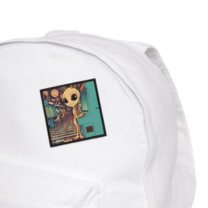 Könnte beinhalten: Ein weißer Rucksack mit einem quadratischen Aufnäher, der einen Alien beim Spielen eines Arcade-Spiels zeigt. Der Aufnäher hat ein Retro-Farbschema mit dem Alien in einem türkisfarbenen Spielautomaten. Der Hintergrund zeigt weitere Spielautomaten.