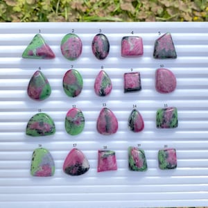 May include: A collection of twenty polished ruby zoisite gemstones, each with unique shapes and patterns. The stones display a mix of green and pink hues, with variations in size and form. The gemstones are arranged on a white surface against a backdrop of green foliage.