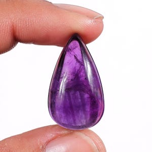 Könnte beinhalten: Ein polierter, tropfenförmiger Amethyst-Edelstein. Der Edelstein zeigt einen tiefvioletten Farbton mit helleren Streifen und inneren Mustern. Der Stein wird zwischen zwei Fingern gehalten, was seine glatte, reflektierende Oberfläche zeigt. Dieser Edelstein eignet sich für die Schmuckherstellung.