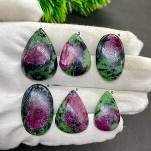 May include: Six polished ruby zoisite gemstones. Each stone features a mix of deep magenta and green hues. The shapes include teardrop and oval forms. The gemstones are displayed on a white surface.