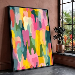 May include: Abstract art print in a black frame. The artwork features a colorful pattern of overlapping shapes in shades of yellow, pink, green, and blue. The painting is displayed against a brown wall near a window with a small potted plant.