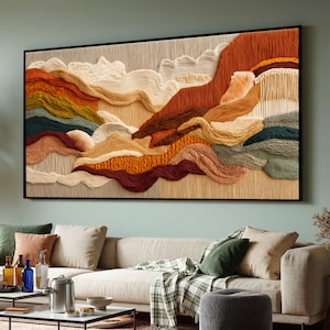 Puede incluir: Una gran obra de arte textil enmarcada que representa un paisaje estilizado. La obra presenta fibras texturizadas y en capas en tonos naranja, marrón, beige y verde, creando una escena de montaña y nubes. La pieza está montada en un marco negro y cuelga sobre un sofá beige.