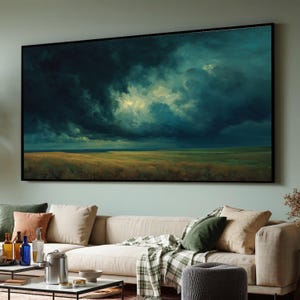 May include: A framed landscape painting depicts a dramatic sky filled with dark, swirling clouds above a field of golden grass. The artwork has a black frame and hangs above a beige sofa in a living room.