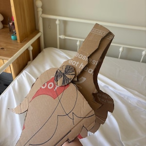 Puede incluir: Un accesorio de cartón hecho a mano con forma de ala o pluma estilizada, con un logotipo rojo y blanco y el texto "easiest eat hea". Los bordes están delineados con un rotulador negro. El accesorio se sostiene contra una sábana blanca.