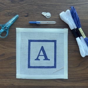 May include: A needlepoint kit with a white canvas featuring a blue square and the letter "A". The kit includes blue scissors, embroidery floss in white and blue, a needle, and a silver thimble. The items are arranged on a wooden surface.