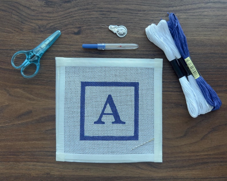 May include: A needlepoint kit with a white canvas featuring a blue square and the letter "A". The kit includes blue scissors, embroidery floss in white and blue, a needle, and a silver thimble. The items are arranged on a wooden surface.