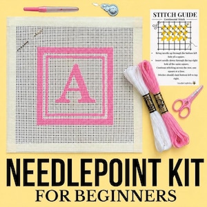 May include: A needlepoint kit featuring a square canvas with a pink letter A, a needle, a pink pen, a silver needle minder, pink scissors, and two skeins of embroidery floss in white and pink, accompanied by a printed stitch guide card.