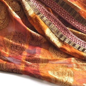 May include: A sheer orange fabric scarf featuring a repeating gold paisley pattern and a plaid base design. The edge is finished with a decorative border consisting of dark brown, metallic gold, and maroon bands with a geometric zigzag motif.