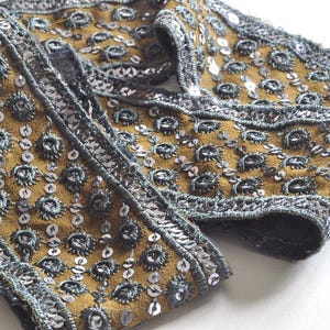 May include: A decorative fabric trim featuring a mustard yellow base with an intricate pattern of dark grey embroidered circular motifs and silver sequins arranged in a grid, finished with a textured silver metallic border along the edges.