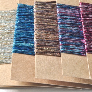 May include: Six rectangular brown cardboard cards wrapped with metallic embroidery thread in various colours including silver, blue, bronze, multicoloured, and deep pink. Each card features a flat, horizontal winding of shiny, reflective thread.