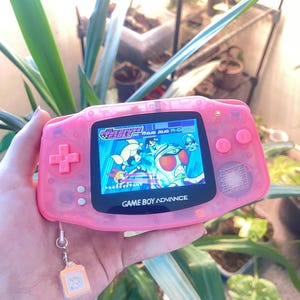 Könnte beinhalten: Eine durchscheinende pinke Game Boy Advance Handheld-Spielkonsole mit einem hintergrundbeleuchteten Bildschirm, der das Spiel The Powerpuff Girls anzeigt. Das Gerät verfügt über ein Steuerkreuz, A- und B-Tasten sowie einen kleinen Anhänger.