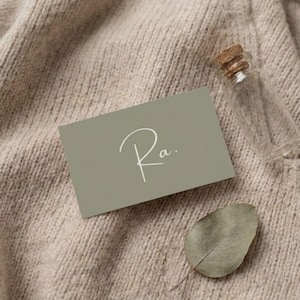 Puede incluir: Una tarjeta de visita verde salvia con la palabra manuscrita "Ra." en escritura blanca, una peque&ntilde;a botella de vidrio transparente con tap&oacute;n de corcho y una hoja seca descansan sobre una superficie de punto beige. El texto "thoughtfully by: GreyMatterHub" est&aacute; en la parte inferior.