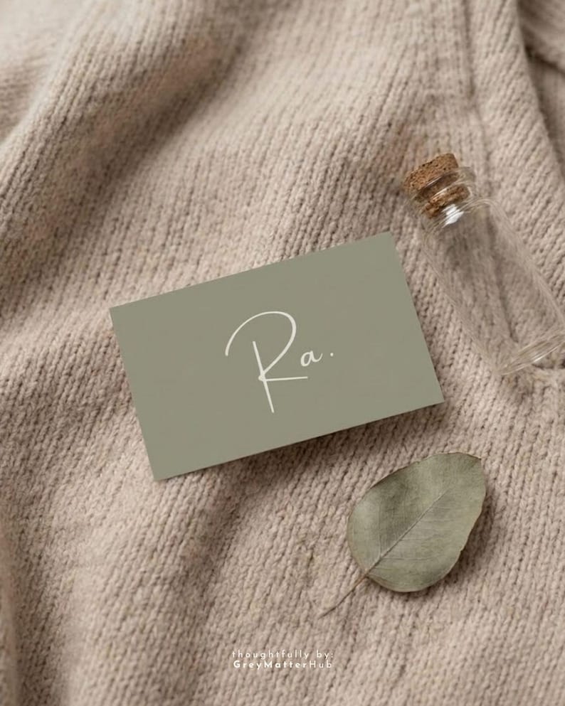 Puede incluir: Una tarjeta de visita verde salvia con la palabra manuscrita "Ra." en escritura blanca, una peque&ntilde;a botella de vidrio transparente con tap&oacute;n de corcho y una hoja seca descansan sobre una superficie de punto beige. El texto "thoughtfully by: GreyMatterHub" est&aacute; en la parte inferior.
