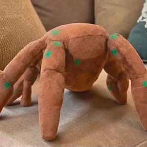 Peut inclure: Un jouet en peluche en forme d'araignée marron avec huit pattes et des points verts. Le jouet est fait d'une matière douce et est posé sur une surface de couleur neutre. Les pattes de l'araignée sont déployées, lui donnant un aspect ludique.