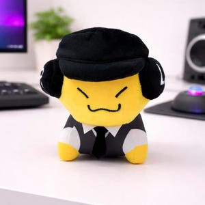 May include: A plush toy character with a yellow face, black hat, and a suit. The toy has a smiling expression and is wearing a black tie. The toy is sitting on a white surface.