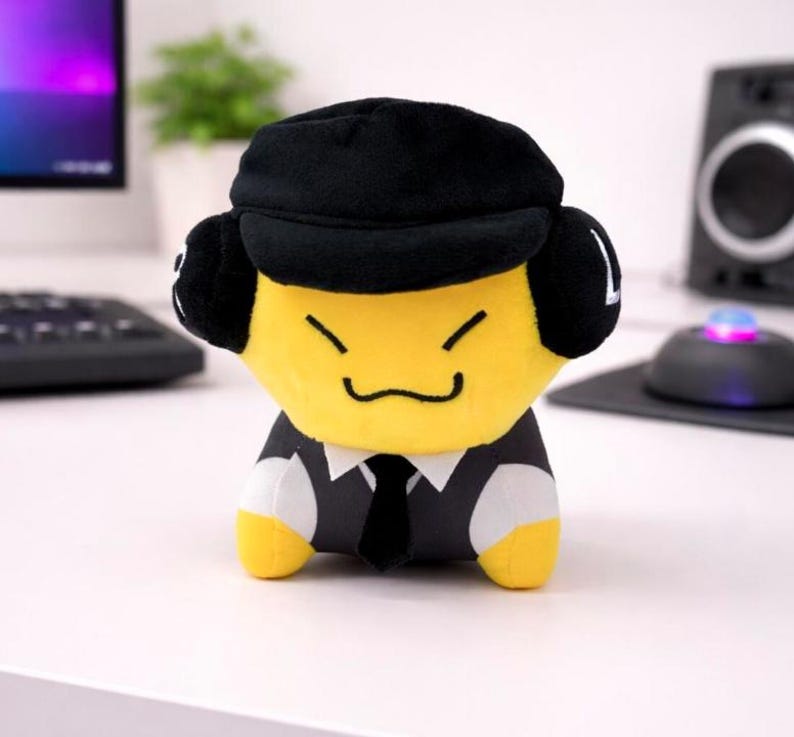 May include: A plush toy character with a yellow face, black hat, and a suit. The toy has a smiling expression and is wearing a black tie. The toy is sitting on a white surface.