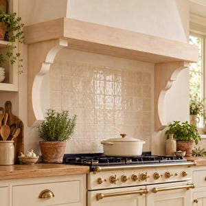 Può includere: Una cappa da cucina con una mensola in legno chiaro sostenuta da due mensole decorative. La cappa è posizionata sopra un piano cottura a gas con manopole in ottone e una pentola color crema, davanti a un paraschizzi di piastrelle beige quadrate.