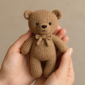 May include: A small, knitted teddy bear in a warm brown colour. The bear has a bow tie, small black eyes, and a stitched nose. The toy is held in open hands, showcasing its soft texture and detailed craftsmanship. A classic children's toy.
