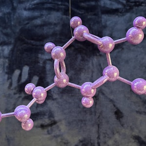 May include: A three-dimensional molecular model structure featuring a series of interconnected spheres and rods. The entire assembly is finished in a light purple color with a noticeable glittery, sparkling texture throughout the surface.